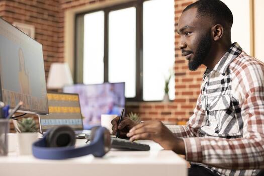 Black male digital artist works from home studio, using stylus and graphics tablet to create and edit portraits on both laptop and desktop screens. Focused illustrator refining visual content remotely photo