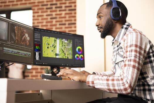 Sound editor working on cinematic content with headphones, creating and synchronizing audio files to montage. Black videographer applying color grading and finetuning transitions while enjoying music. photo