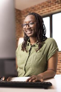 Portrait of cheerful freelancer typing on computer keyboard while reviewing business strategies. Happy african american woman using desktop pc in comfort of home, working remotely on office reports. photo