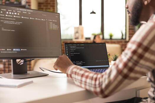 African american tech freelancer programming secure systems and reviewing coding scripts in apartment. Black male IT specialist working with multiple devices in modern home office setup. photo