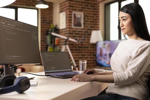 Determined asian woman seated at home with laptop and desktop monitor, confidently troubleshooting coding scripts. Remote worker focused on debugging software in cozy brick wall apartment. photo