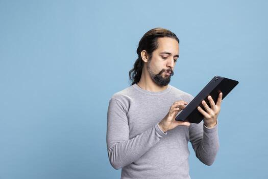 casual y confidente joven hombre utilizando un tableta en frente de el cámara, resolviendo independiente Tareas en línea. enfocado moderno persona lo hace digital trabajo o social redes en contra azul antecedentes. foto