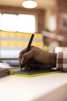 Close up of african american hand using stylus and graphic tablet to retouch images on laptop. Black freelancer utilizing efficient digital photo editing setup in creative home workspace.
