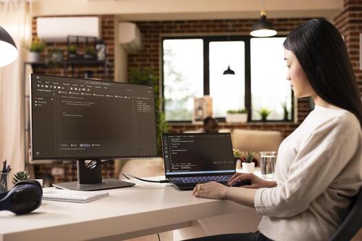 Focused female developer programming on laptop in home office. Asian woman building software and coding website remotely from modern apartment using digital tools for web development. photo