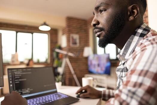 African American man sits at desk in modern apartment, concentrating on laptop and computer screens. Serious expression shows deep focus on code development and programming work. photo