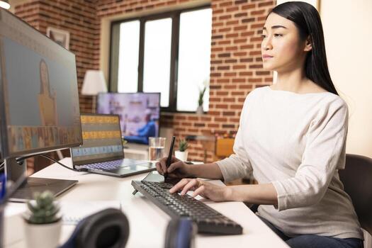 Asian graphic designer working with digital drawing pad, retouching images and enhancing design elements. Skilled photo editor perfecting visual details on computer screen in creative home studio.