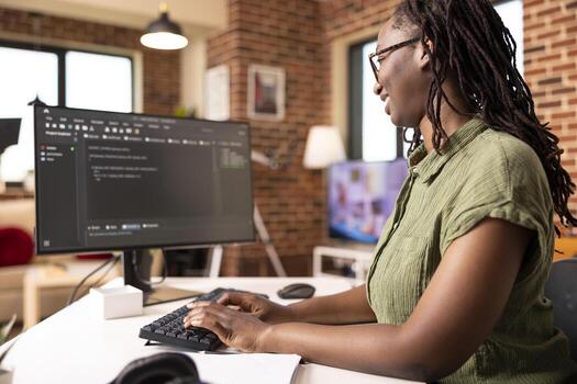Black female software developer codes securely from home office, using computer to review programming scripts. African american IT freelancer working on tech solutions in modern apartment. photo