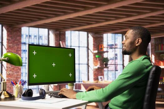 Happy man using chroma key PC in apartment to write emails. Smiling remote worker typing on mockup computer keyboard at home office desk, composing documents, doing work tasks photo