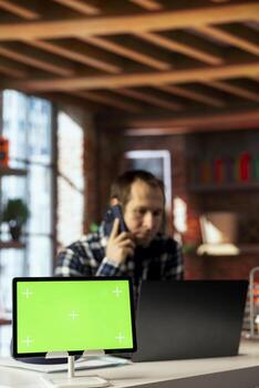 Close up of green screen tablet next to man answering phone call while checking emails. Focus on mockup device in front of worker in blurry background doing telephone call with friend at work photo