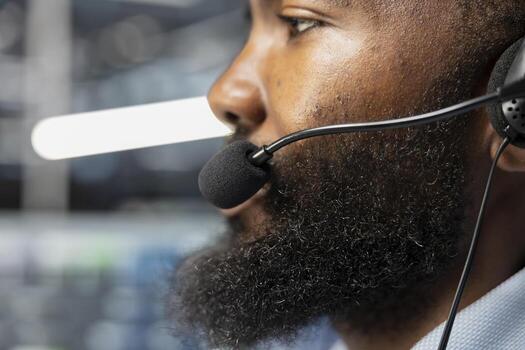Close up of data center worker in remote call providing customer support, troubleshooting damages. African american man in server hub talking with clients, using microphone photo