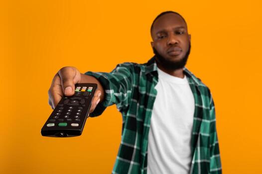 Bored African American Man Switching Channels Pointing Television Controller To Camera Watching Boring TV Program Posing On Yellow Studio Background. Discontented Televiewer. Selective Focus photo
