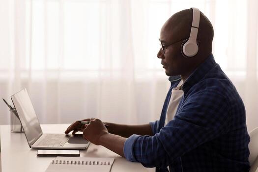 Distant work. Black mature freelancer working with laptop from home office. Man using computer for online project, typing on keyboard, sitting at desk near window, copy space, side view photo