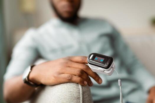 Unrecognizable African Guy With Pulse Oximeter Clip On Hand Measuring Oxygen Saturation Level Monitoring Ox And Pulse Rate At Home. Pulseoxymetry Medical Device Concept. Cropped, Selective Focus photo