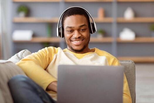 Closeup of happy black guy playing games on laptop while reclining on couch at home, using wireless headset. smiling and looking at computer screen, copy space. Online gaming concept photo