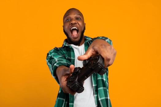 Computer Gaming. Excited African Man Playing Holding Game Controller Having Fun Standing Over Yellow Studio Background, Smiling To Camera. New Videogame Advertisement Concept. Selective Focus photo