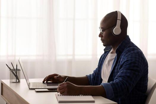 Mature african american freelancer working remotely with laptop at home office. Man using computer for online project, typing on keyboard, sitting at desk near window, copy space photo