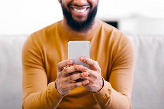 Unrecognizable African Man Using Smartphone Testing New Mobile App, Texting And Browsing Internet Sitting On Couch At Home. Mobile Application And Communication Concept. Cropped, Focus On Phone photo