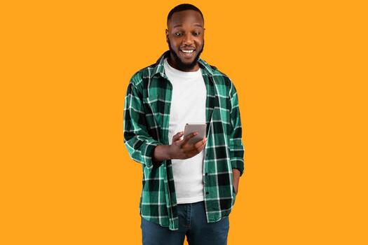 Cheerful Black Man Using Smartphone Browsing Internet And Texting Standing Over Yellow Studio Background. New Mobile Application For Cellphone Advertisement Concept photo