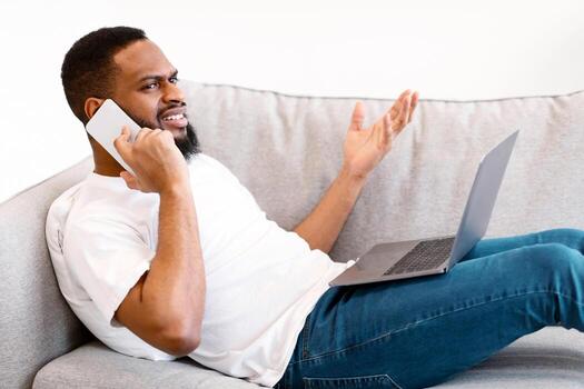 Side View Of Discontented African Guy Talking On Cellphone Using Laptop And Having Problem With Internet Connection Lying On Sofa At Home. Displeased Customer Concept photo