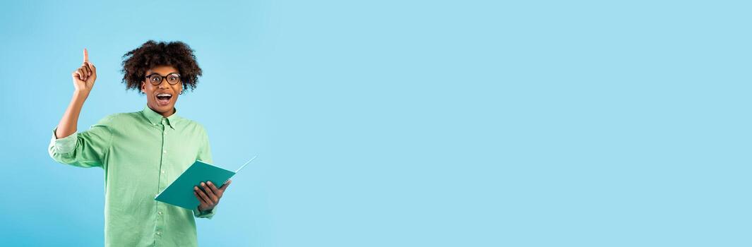 A student holds a book while raising a finger in a thoughtful gesture, showcasing an expression of inspiration. The bright background provides ample space for text or messaging. photo