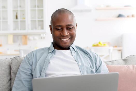 gratis tiempo. contento medio Envejecido negro hombre utilizando ordenador portátil mientras descansando en sofá a hogar. calvo maduro usuario surf en Internet o chateando en línea en computadora, relajante después trabajo foto