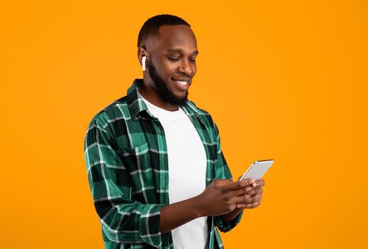 Music Application. African Man Using Phone Wearing Earbuds And Listening To Song Online Standing Over Yellow Studio Background. Smartphone User Browsing Internet On Cellphone photo