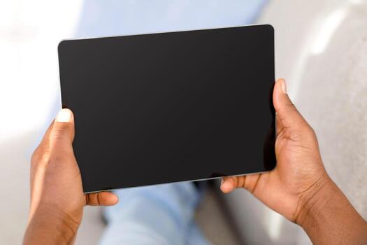 Unrecognizable black man using digital tablet with black blank screen, mockup for application or website advertisement, closeup, crop. Hands holding device with empty space photo