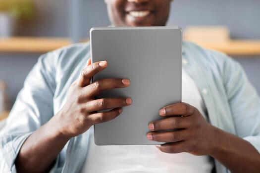 Free time concept. Black man using digital tablet, reading news or browsing internet, enjoying weekend and resting on the couch at home, selective focus. Gadget lifestyle photo