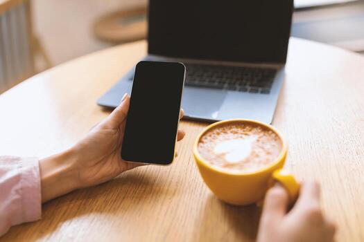irreconocible mujer utilizando Teléfono móvil con negro pantalla mientras Bebiendo café a cafetería, Bosquejo para tu diseño. recortado ver dama utilizando teléfono con espacio para móvil aplicación o sitio web, ordenador portátil en antecedentes foto