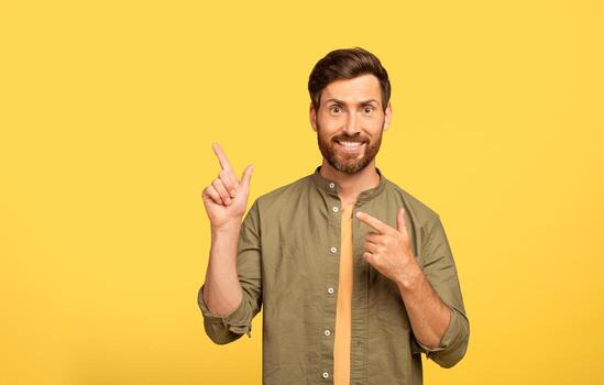 A middle aged man with a beard appears cheerful while pointing with both hands at copy space. He is dressed casually in an olive green shirt, standing in front of a bright yellow backdrop. photo