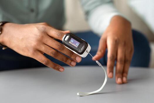 Unrecognizable African Man Using Pulse-Oximeter Checking Oxygen Saturation And Pulse Rate Sitting At Home. Pulseoxymetry Medical Device On Hand Measuring Ox Level. Cropped, Selective Focus photo