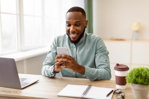 Great Business App. Happy African Businessman Using Phone Reading Good News Message Sitting At Workplace In Modern Office. Entrepreneur Texting Networking Online Via Cellphone Application photo