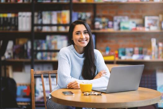 Happy latin lady resting in cafe and using laptop computer, smiling at camera, empty space. Happy freelancer working distantly, surfing internet, sending email or chatting with clients photo