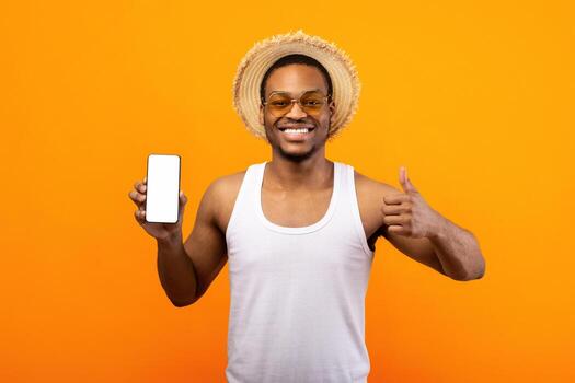 Check out this new mobile application. Happy African American guy showing cellphone with mockup for website or app, gesturing thumb up on yellow studio background photo