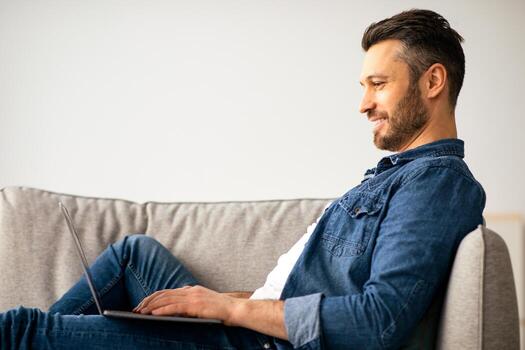 Smiling middle-aged man using modern laptop, having part-time job at home, copy space. Happy bearded man watching movie or chatting on notebook, resting on sofa in living room, side view photo