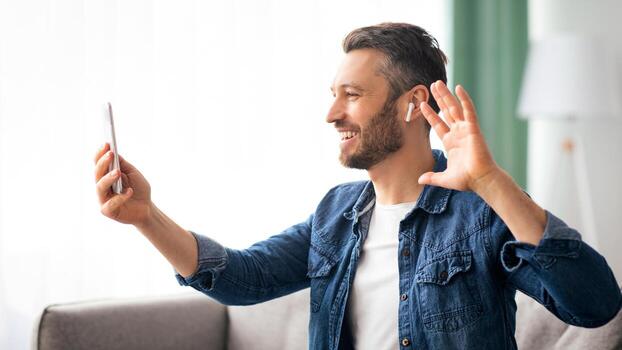 Cheerful bearded man waving at mobile phone while resting on couch at home, side view, copy space. Happy middle-aged man having conference with friends or relatives, using smartphone photo