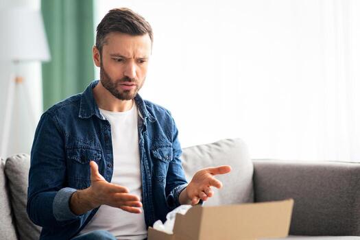 Disappointed middle-aged man customer checking mail box, sitting on sofa at home, copy space. Angry man opening delivery, checking order, having negative face expression. Delivery concept photo