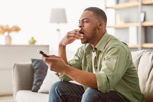 Bored Black Millennial Guy Yawning Watching Boring TV And Switching Channels With Remote Controller Sitting On Couch In Living Room At Home. Bad Television Programming Concept. Side View photo