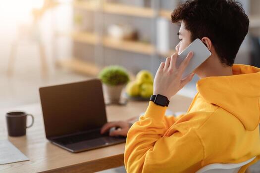 Over the shoulder view of asian man having conversation on mobile phone, using laptop with blank screen, copy space. Male freelancer talking with clients on smartphone while using pc at desk photo