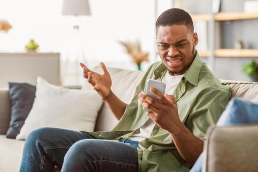 problema con teléfono. disgustado negro hombre utilizando teléfono inteligente teniendo problema con conexión o móvil aplicación sentado en sofá a hogar. descontento Teléfono móvil usuario leyendo negativo mensaje o Noticias foto