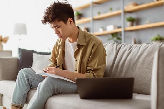 Focused asian man taking notes while working on laptop computer at home office, sitting on sofa. Freelancer noting down information from internet, planning working schedule, signing papers photo