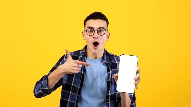 Excited young guy pointing at smartphone with blank screen on orange studio background, space for app or website design. Shocked millennial man showing mobile device with mockup for your ad photo