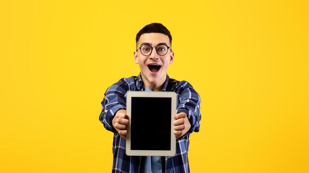 Overjoyed millennial man showing tablet computer with mockup for website or app design on orange studio background. Excited young guy with touch pad, space for your ad photo