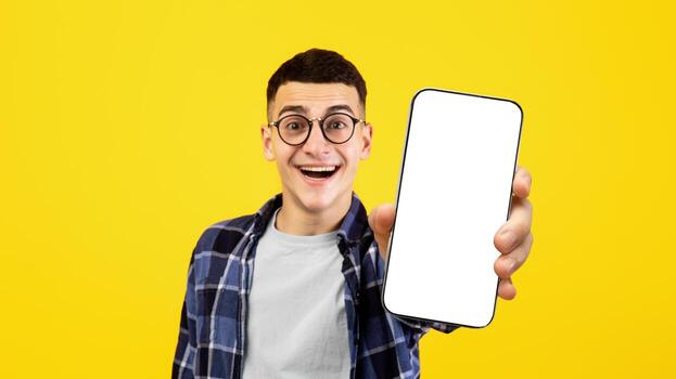Happy young man showing mobile phone with empty screen on orange studio background, mockup for mobile app or website design. Positive guy with blank smartphone, selective focus photo