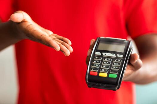 Digital Contactless Transaction. Cropped closeup view of African American man in red t-shirt uniform holding and showing POS machine, selective focus on payment dataphone terminal, blurred background photo