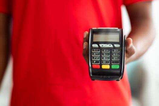 Cropped close up view of African American man in red uniform holding POS machine, selective focus on payment terminal, blurred background. Digital contactless transaction photo
