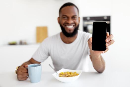Closeup of bearded black male holding smartphone with empty screen in hand, showing device close to camera, sitting at dining table with cereal and coffee, blank space, mock up, blurred background photo