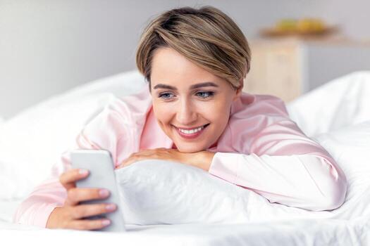 Mobile Phone User. Closeup portrait of happy woman lying in bed on white pillow, smiling reading text message from someone, holding and using smartphone, chatting with lover or browsing social media photo