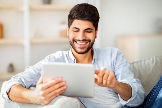 Happy arab man using digital tablet, shopping online, browsing internet or working remotely on tab computer, sitting on sofa and enjoying domestic leisure and modern technologies photo