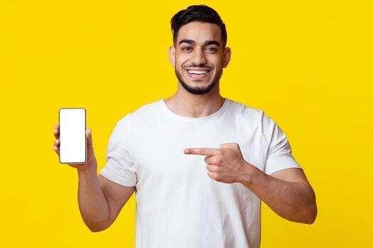 Happy arabic millennial man pointing at brand new smartphone with empty screen in his hand and smiling, recommending newest mobile application, mockup, copy space, yellow background photo